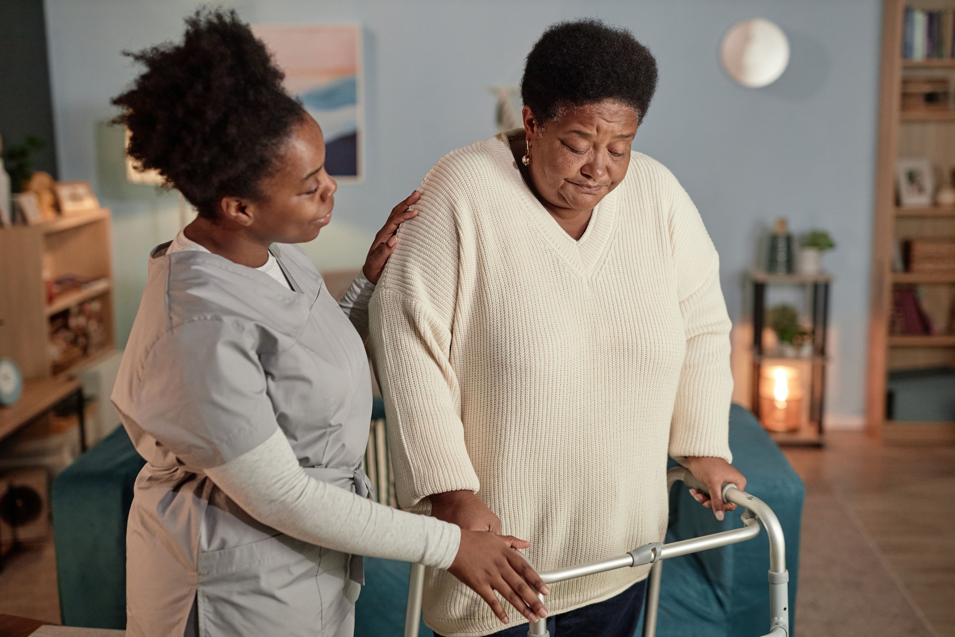 Black Female Caregiver Supporting Senior Woman Walking with Frame while Helping around House